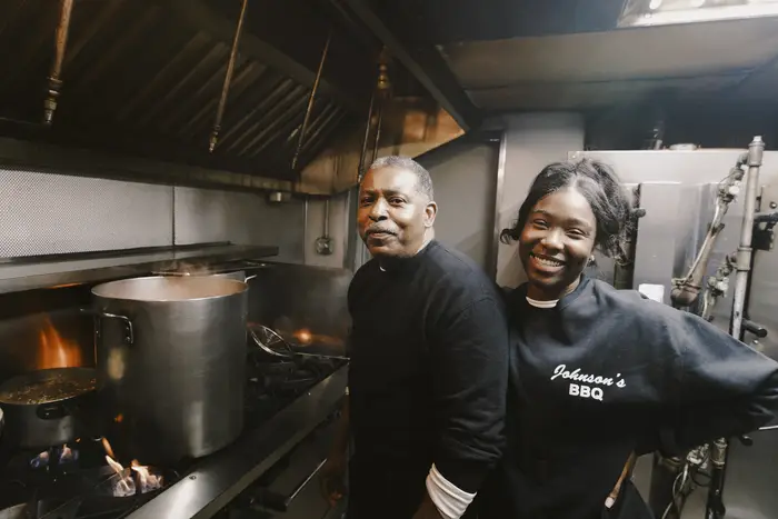 Two chefs, smiling at the camera, in a kitchen, standing next to a large pot.